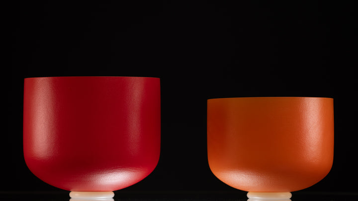 Two frosted crystal singing bowls on stands, one red and one orange, against a black background.