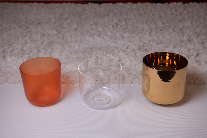 Three crystal singing bowls in different sizes with a gold-colored exterior, an orange-colored bowl, and a clear quartz singing bowl, displayed on a white surface.