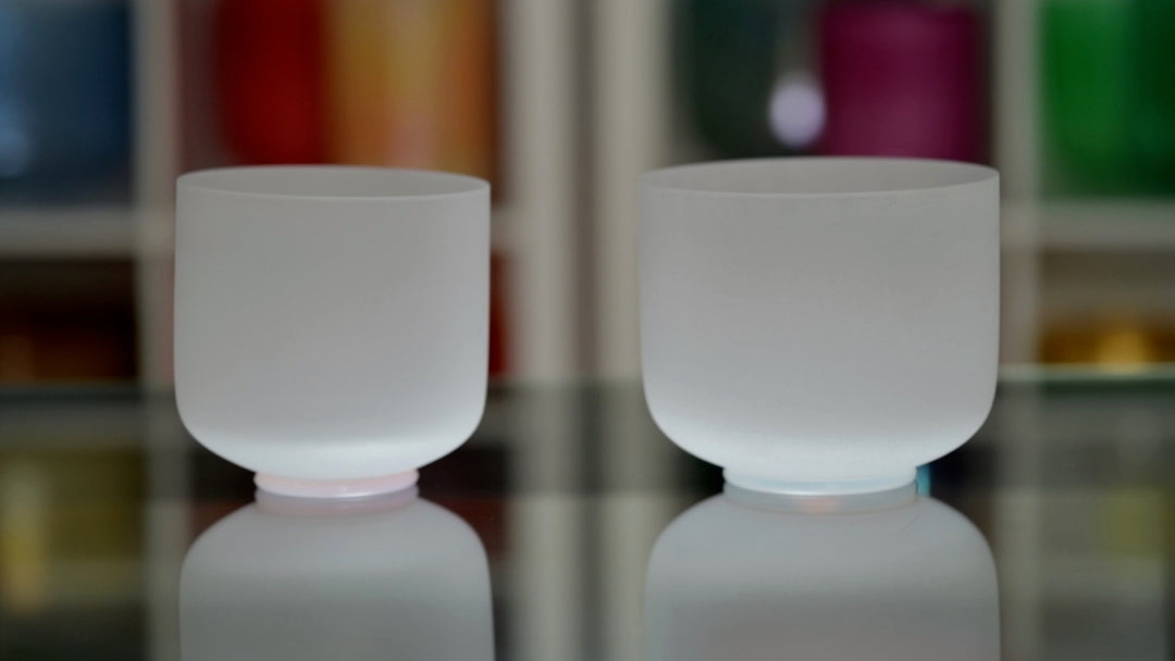 Two white frosted crystal singing bowls on a reflective surface, with one slightly larger than the other.