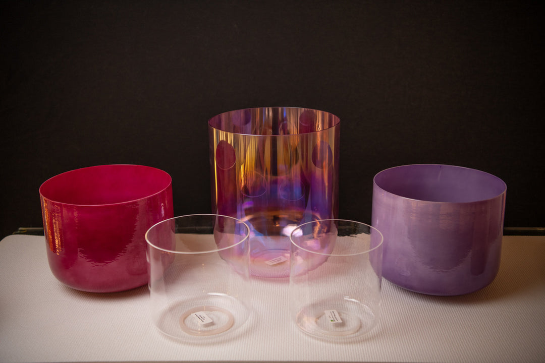 A set of five different sized crystal singing bowls, with varying colors and a clear quartz bowl, displayed on a white surface with a dark background.