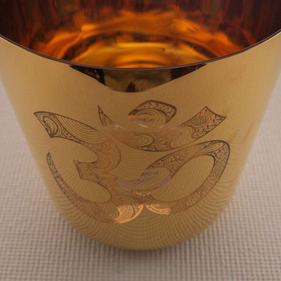 A 9-inch gold crystal singing bowl with a sacred om symbol engraved on it, placed on a textured surface.