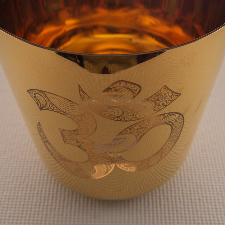 A 9-inch gold crystal singing bowl with a sacred om symbol engraved on it, placed on a textured surface.