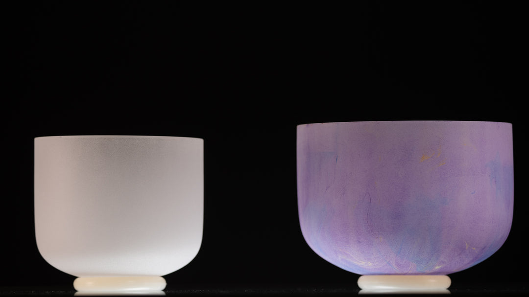 Two frosted crystal singing bowls, one white and one light purple, displayed side by side against a dark background.