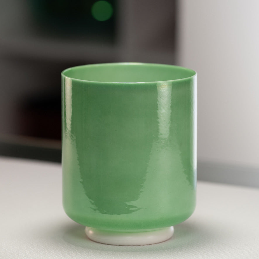 A green crystal singing bowl with a smooth surface, placed on a white surface, with a blurred background.