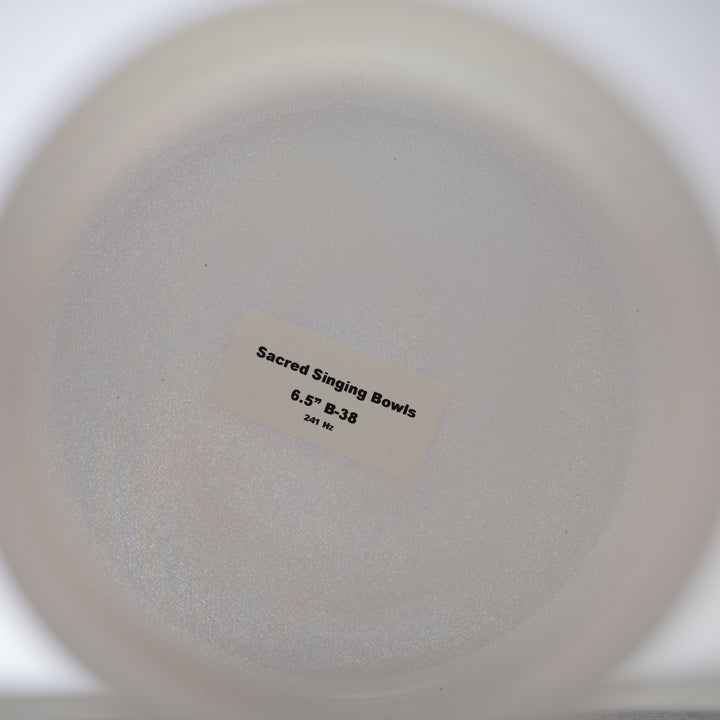 A white 6.5" B-38 Majestic Moon Crystal Singing Bowl with a label that reads 'Sacred Singing Bowls', placed on a clear surface.