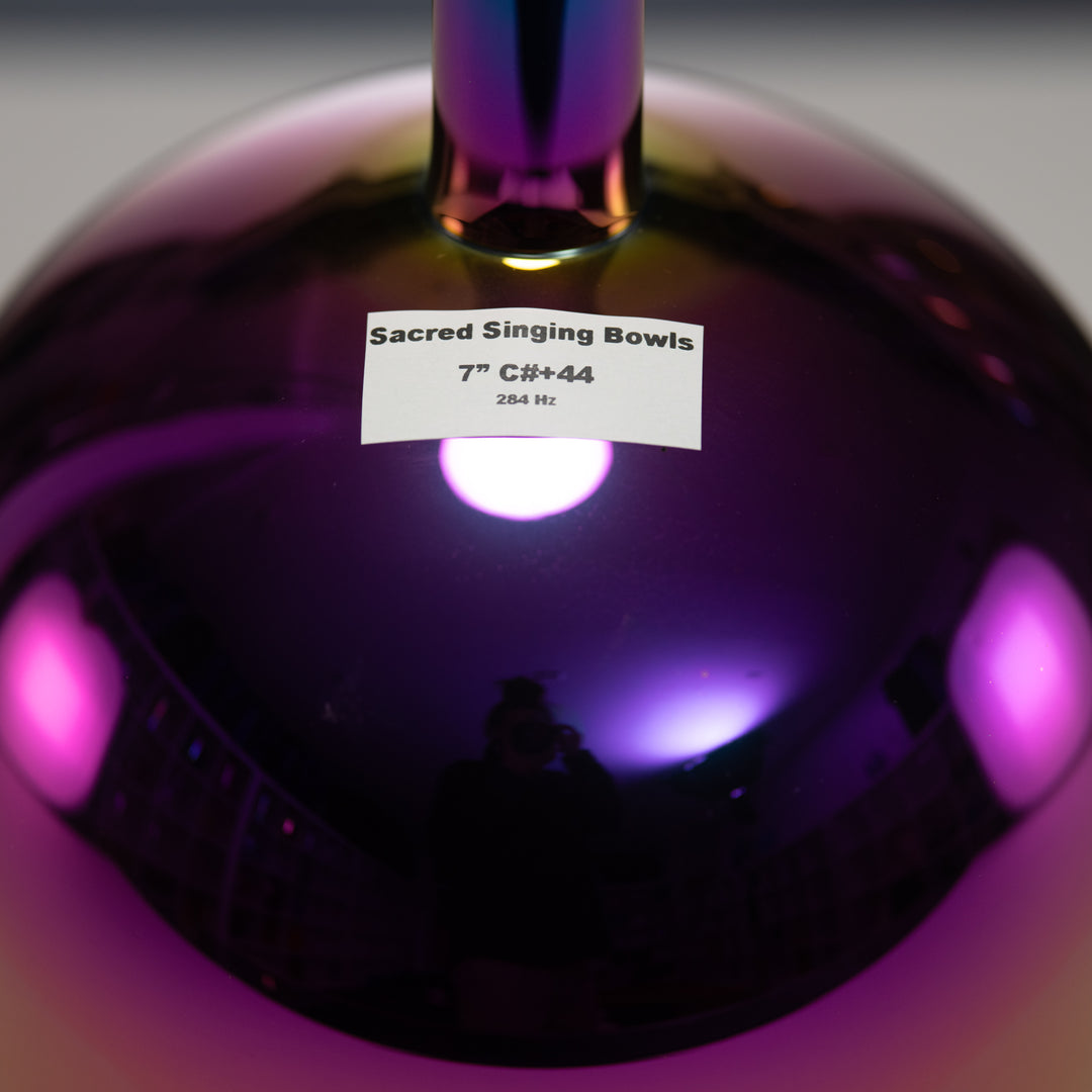 A 7-inch Celestial Rainbow Handheld Singing Bowl with a label indicating it's from Sacred Singing Bowls, 7" C#44. The bowl is shiny with a reflective surface showing iridescent colors.