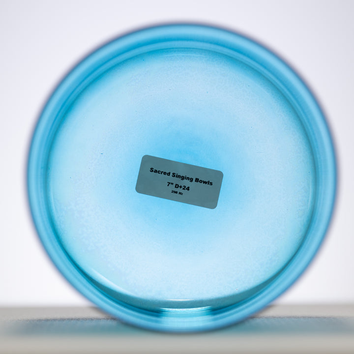 A 7 inch D+24 Caribbean Blue Crystal Singing Bowl with a label in the center that reads 'Sacred Singing Bowls 7" D+24 Hz'. The bowl is placed on a blurred background.