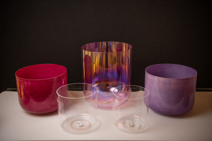 A set of five different sized crystal singing bowls, with varying colors and a clear quartz bowl, displayed on a white surface with a dark background.